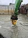 Baggerarm mit gelbem Werkzeug taucht in trübes Wasser einer Baugrube ein