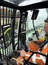 Baggerführer steuert in moderner Kabine mit mehreren Bildschirmen eine Baumaschine an einem Hafen.