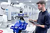 CAPTION: Ein Roboterarm arbeitet in einer hellen Produktionshalle an einem blauen Bauteil, während ein Mitarbeiter ein Tablet zur Steuerung hält.